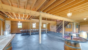 A spacious indoor area with wooden beams, a high ceiling, and a central table with chairs. There are windows allowing natural light, and a staircase leading to an upper level. - Lakefront Gem: Hot Tub & Game Room in Hancock (Hancock)