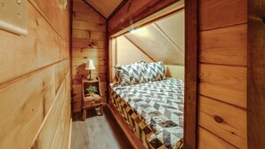 A wooden cabin bedroom with a sloped ceiling, a bed with patterned bedding, a nightstand with a lamp, and a small plant. - Lakefront Gem: Hot Tub & Game Room in Hancock (Hancock)
