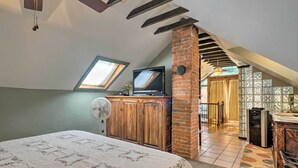 A bedroom with a bed, a TV, a fan, a brick wall, and a view of a kitchen and dining area. - Historic Manitou Springs Victorian: Walk Downtown! (Manitou Springs)