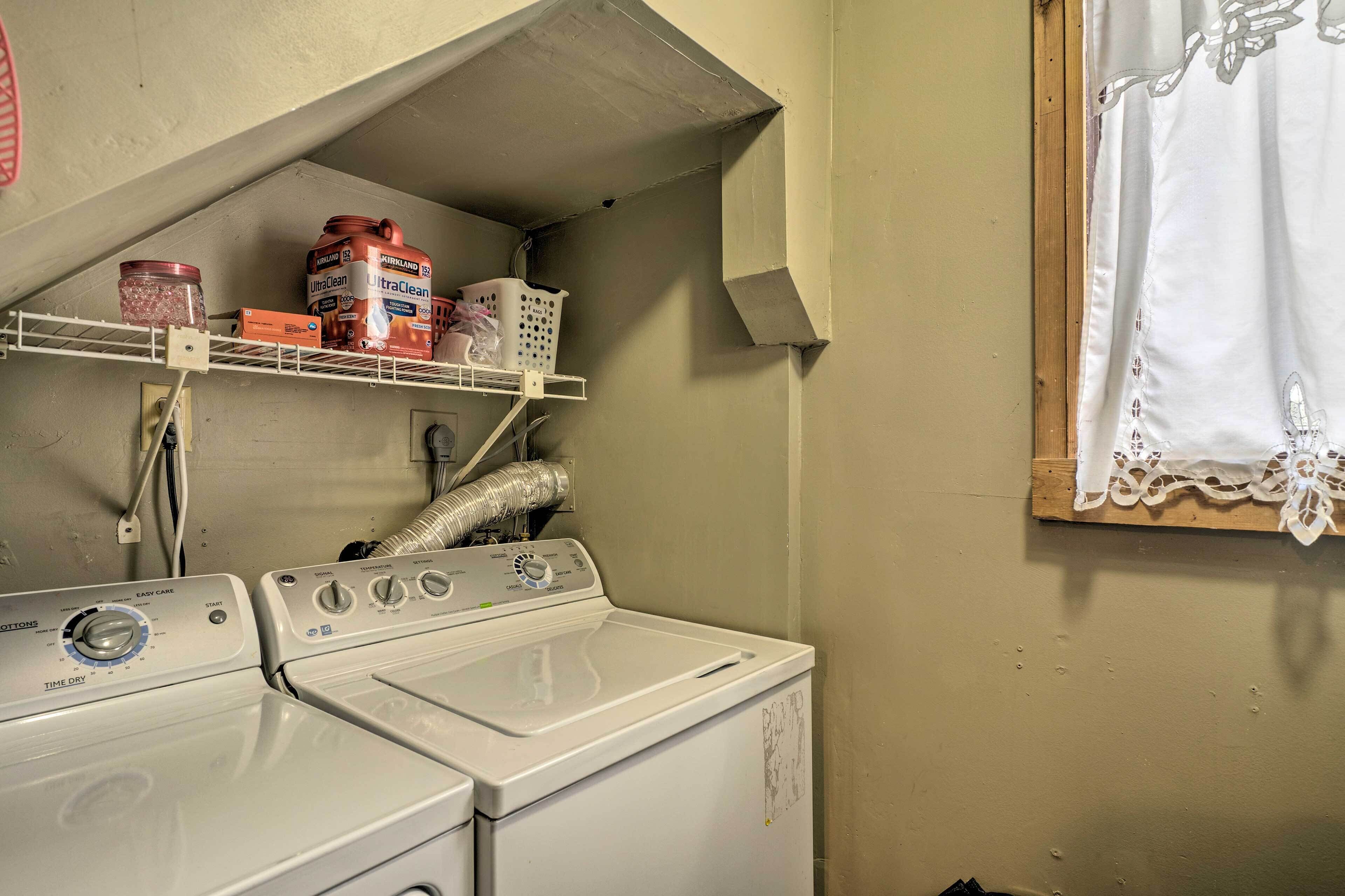 A laundry area with a washing machine, a shelf with laundry supplies, and a window with a curtain.