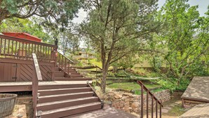 A wooden deck with steps leading to a covered area, surrounded by trees and a stone wall. - Historic Manitou Springs Victorian: Walk Downtown! (Manitou Springs)
