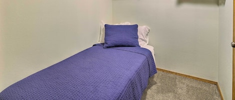 A small, neatly organized bedroom with a single bed, a wooden shelf above holding folded clothes and a plastic bag, and a door to the right.