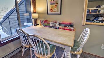 Un coin repas avec une table, deux chaises blanches et une fenêtre avec des stores. On peut voir des jeux de société sur la table.