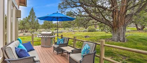 A wooden deck with wicker furniture, a blue umbrella, and a large tree in the background.