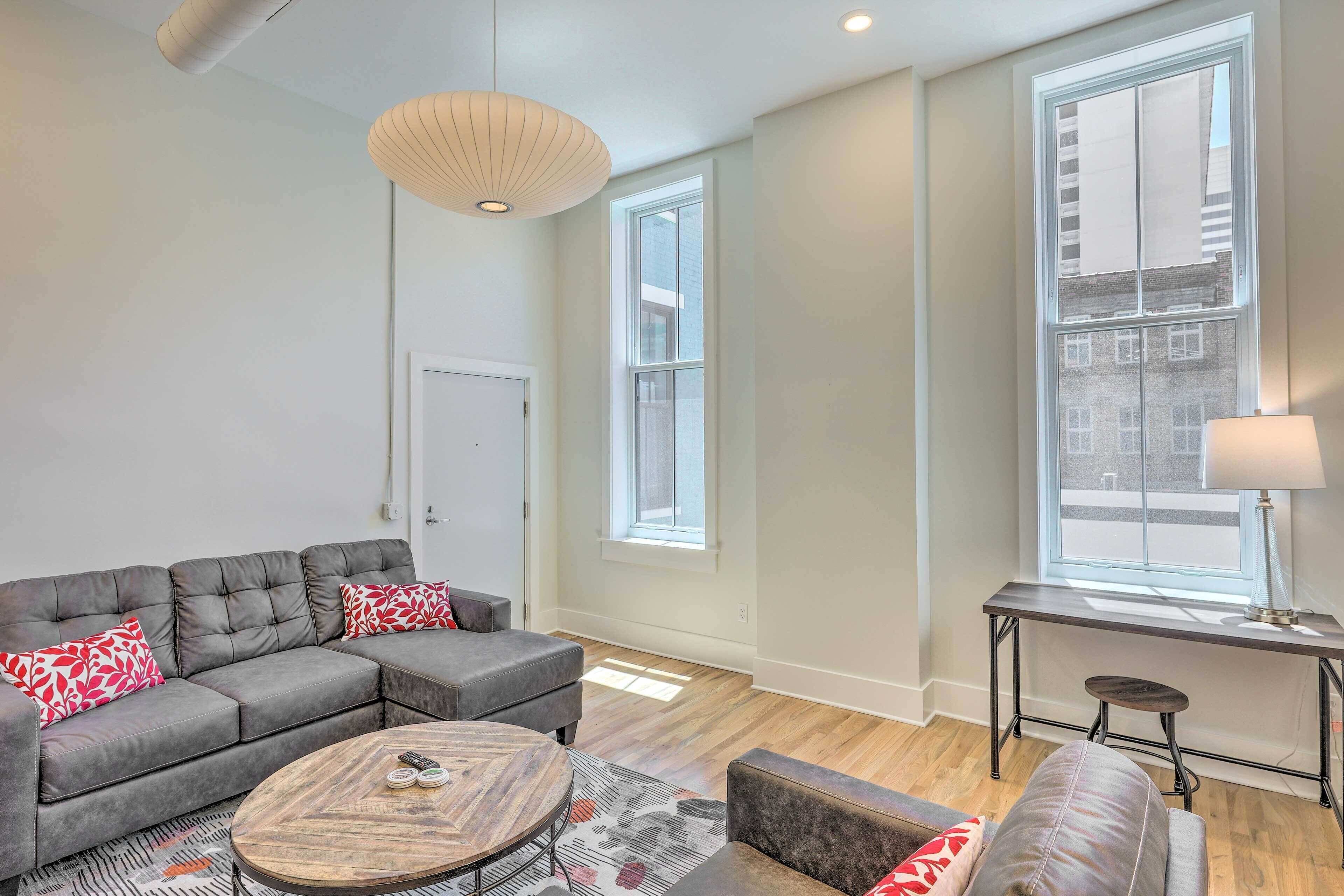 A modern living room with a grey sectional sofa, a round coffee table, and a small stool. There are two windows providing natural light and a view of the city.