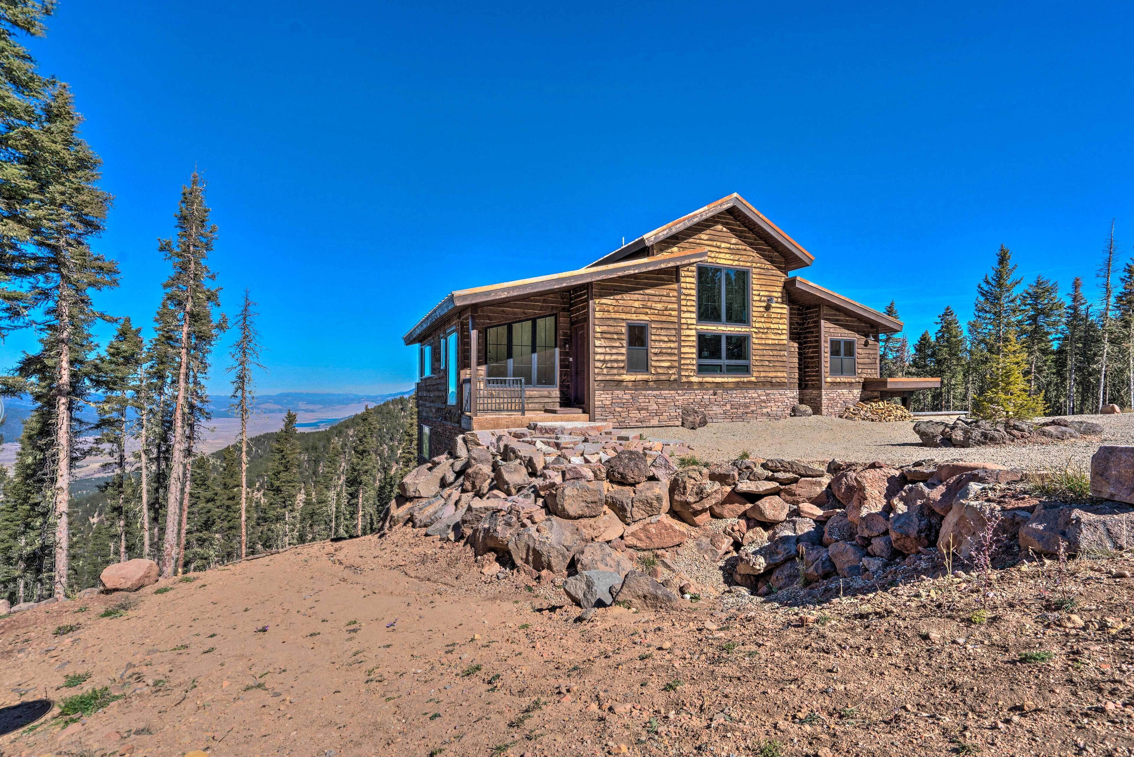 A log cabin with a stone foundation, large windows, and a view of the surrounding forest and mountains.