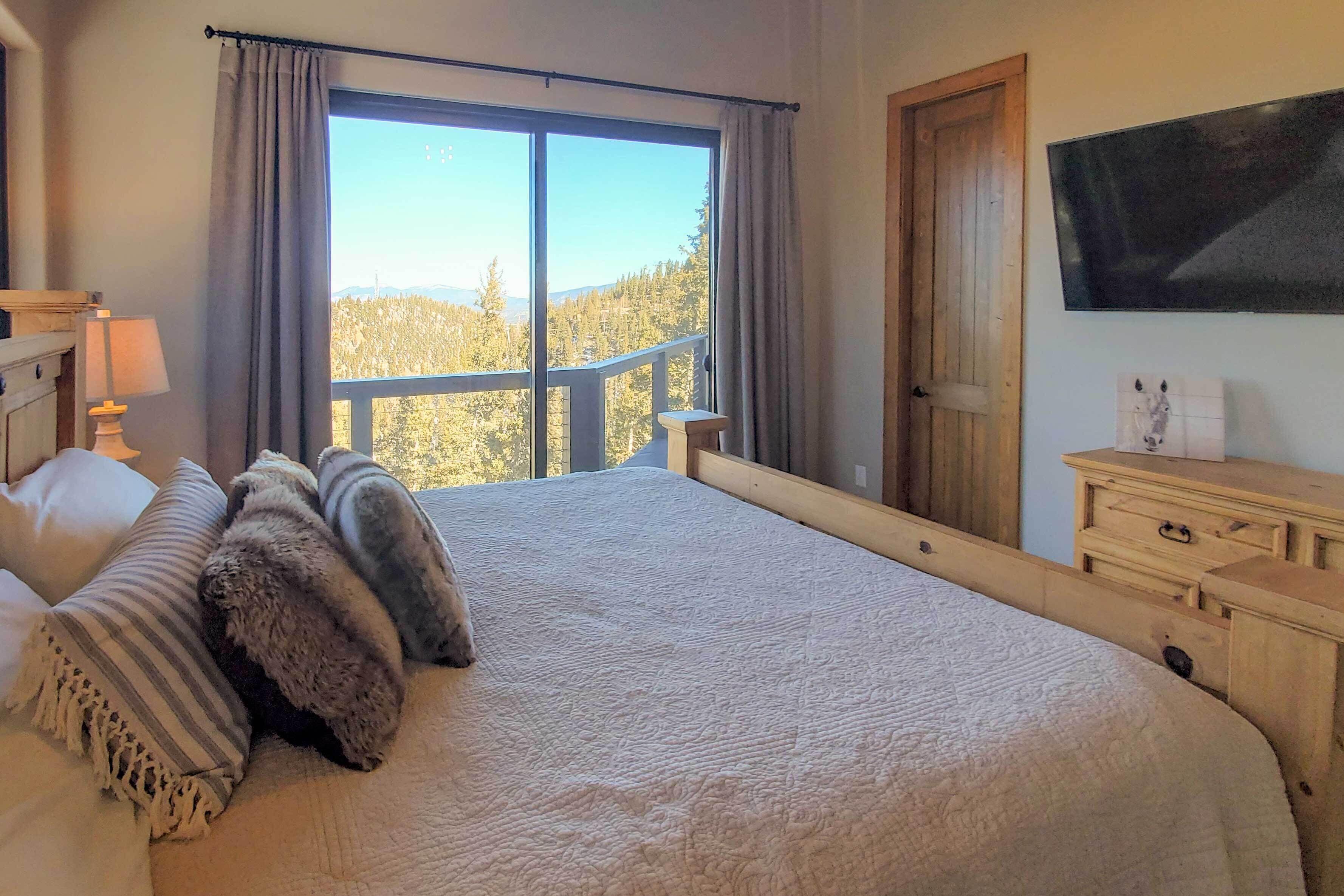 A bedroom with a large bed, a wooden dresser, a TV on the wall, and a window with a view of trees and mountains.