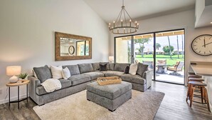 Ein modernes Wohnzimmer mit einem Ecksofa, einem runden Hocker, einer Kristallleuchtre und einem großen Fenster mit Blick auf einen Golfplatz.
