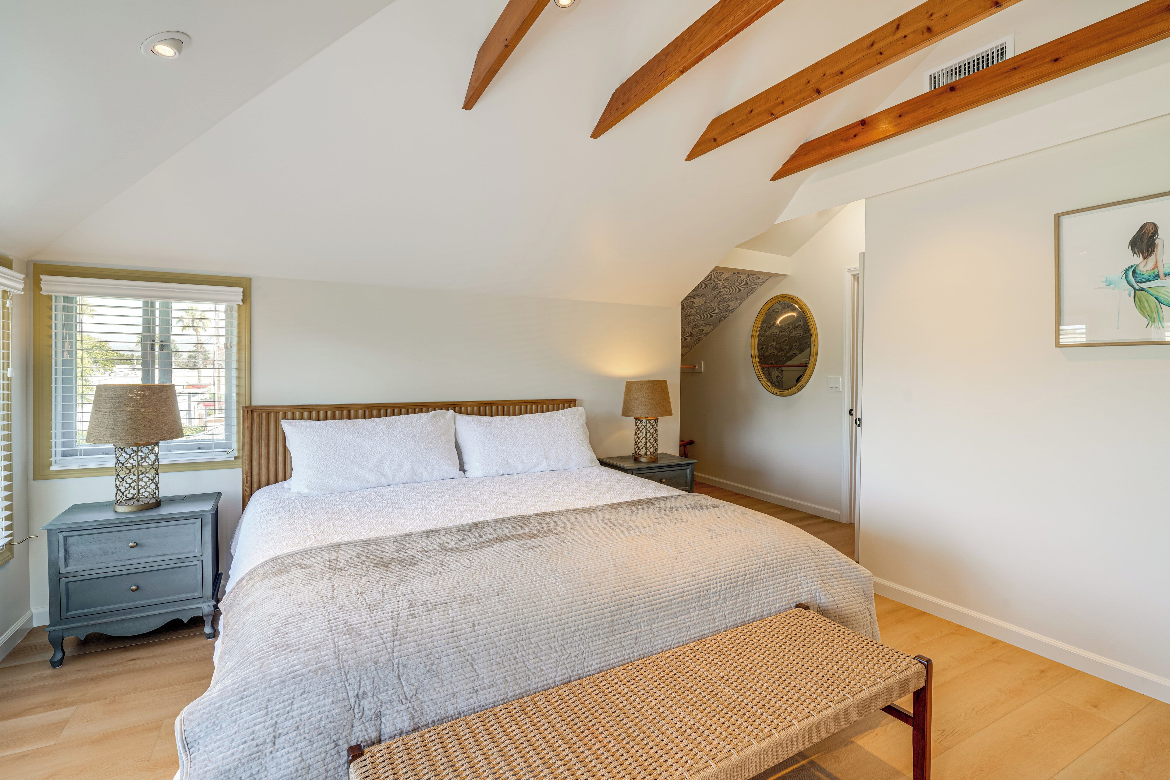 A bedroom with a large bed, wooden beams, a nightstand with a lamp, a bench, and a wall with a framed picture.