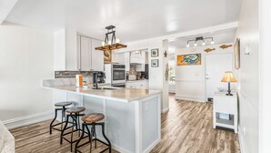A modern kitchen with white cabinetry, a central island with stools, a hanging light fixture, and a view into the dining area. - Oceanfront St Helena Island Condo w/ Beach Access! (Harbor Island)