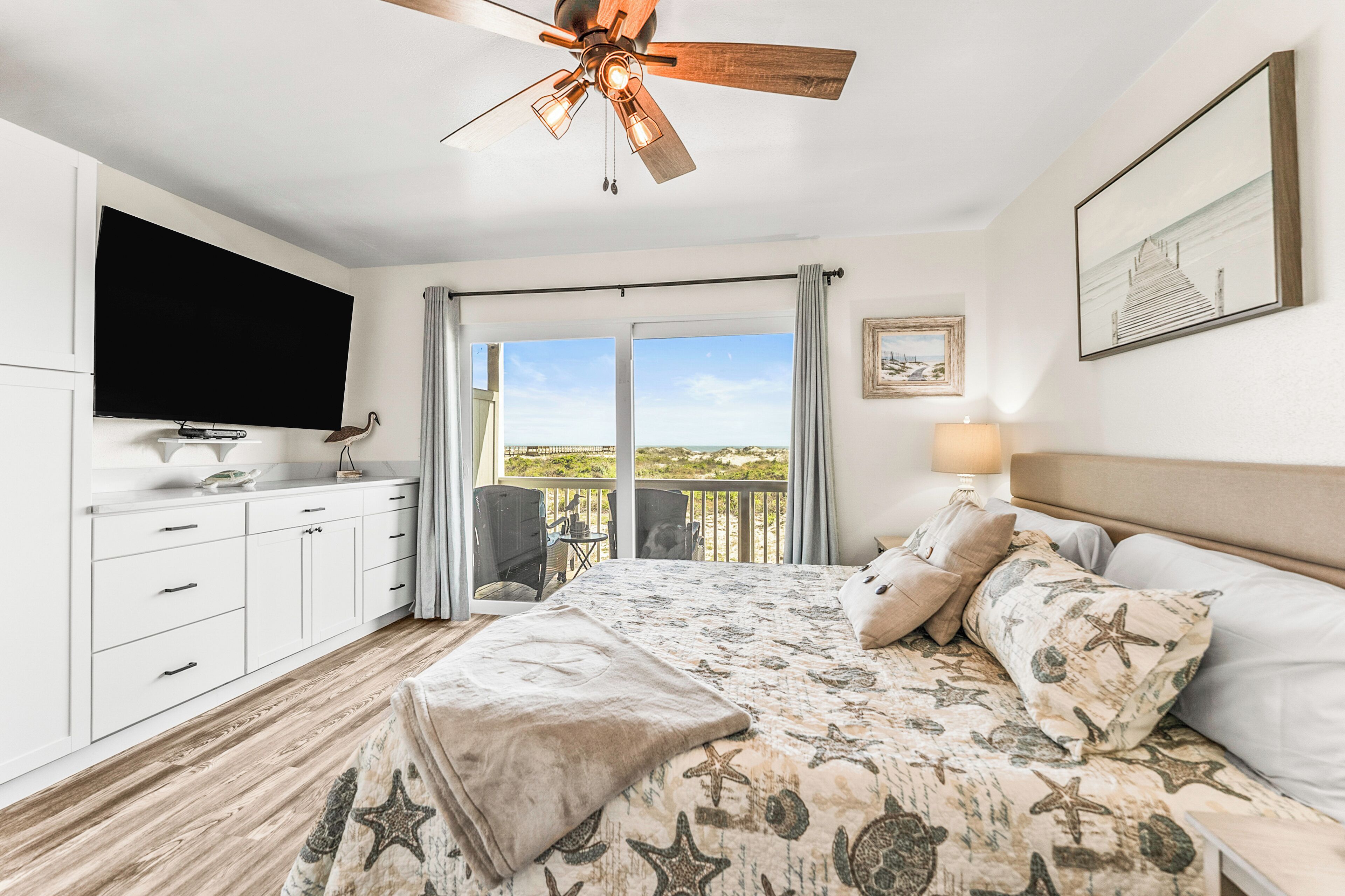 A bedroom with a bed, a ceiling fan, a TV, a balcony with a view, and a painting on the wall.