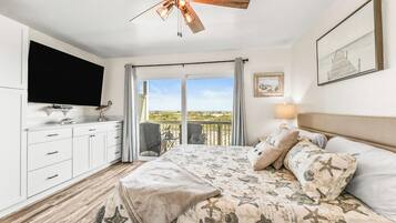 A bedroom with a bed, a ceiling fan, a TV, a balcony with a view, and a painting on the wall.