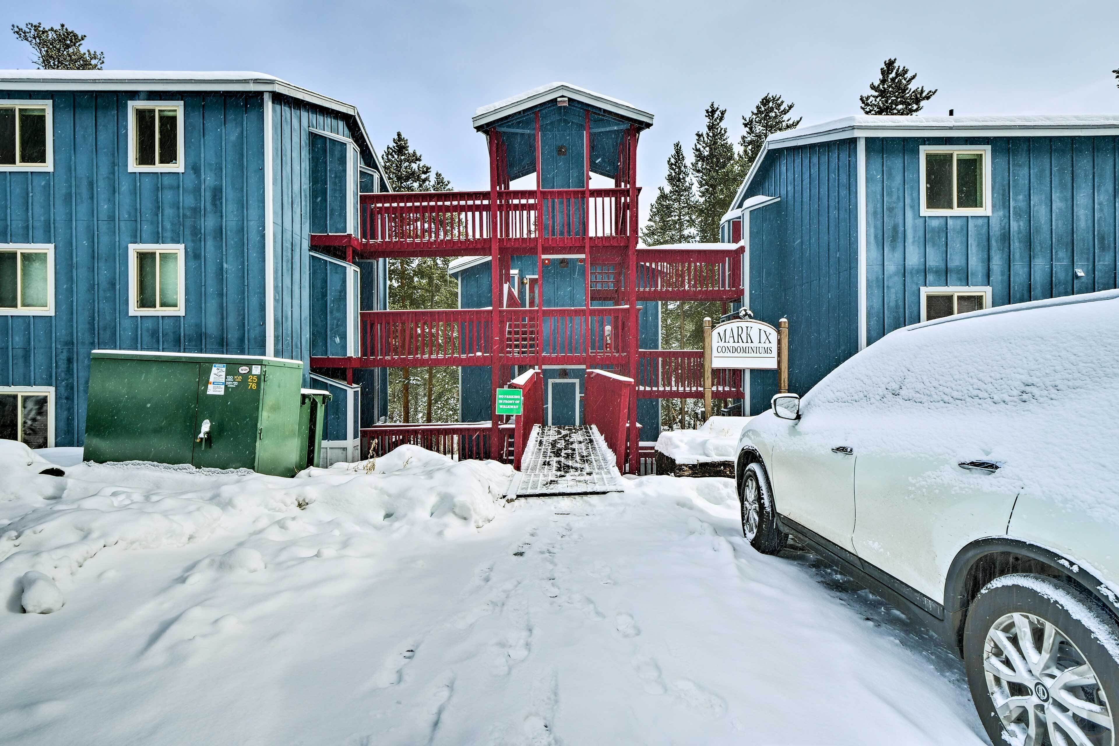 Un parking enneigé avec une voiture blanche, un bâtiment rouge et bleu doté d’un balcon, et un panneau indiquant la présence de copropriétés.