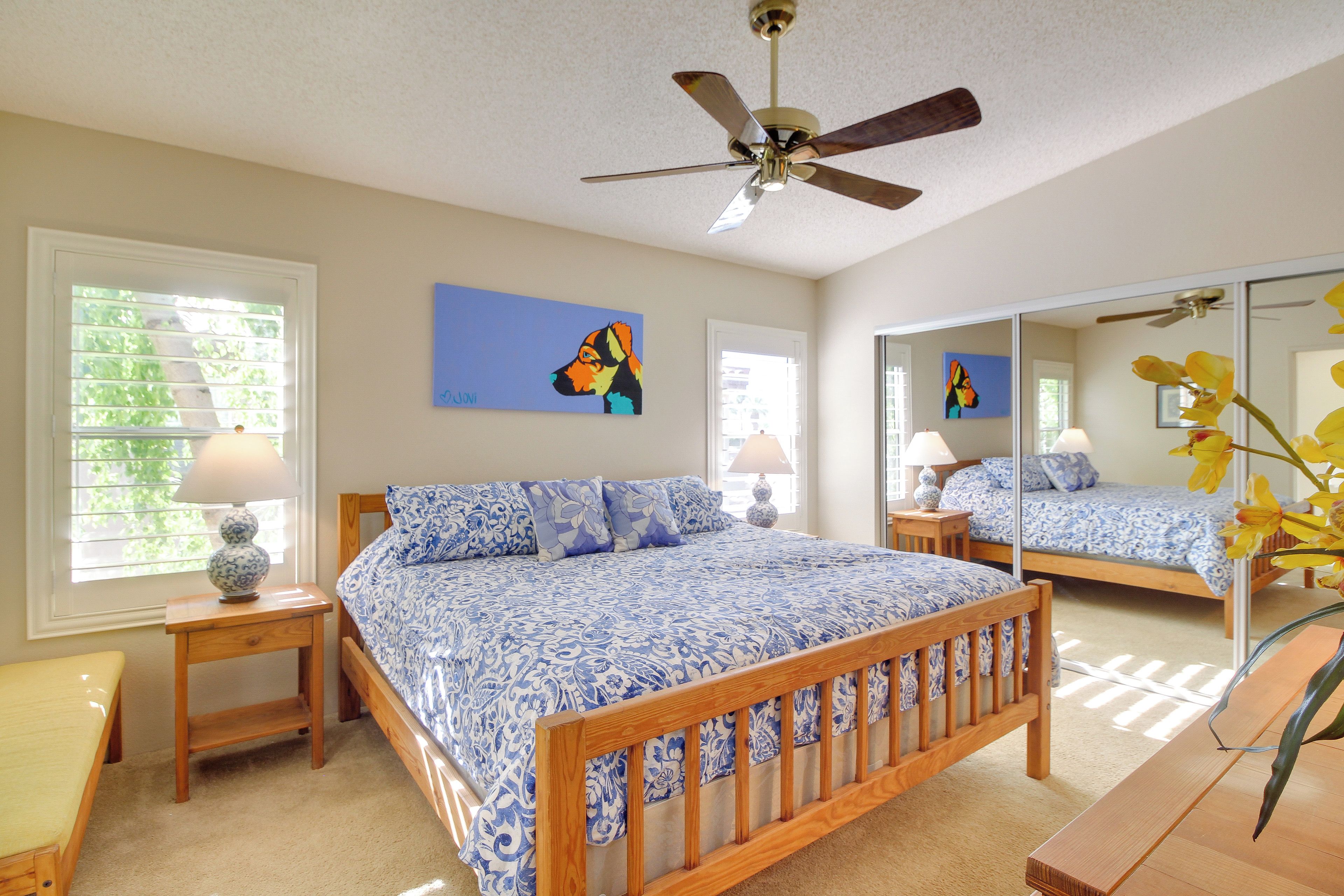 A bedroom with a wooden bed, a ceiling fan, a window with blinds, and a mirror reflecting the room.