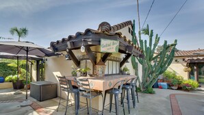 Een terras buiten met een tafel en stoelen, een bordje met 'Cocktail Hour' erop, en een grote cactus op de achtergrond.