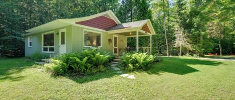 A small, single-story house with a covered porch, surrounded by lush greenery and trees.