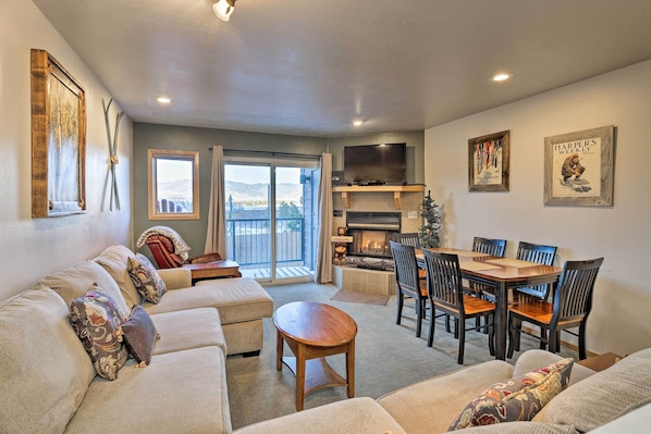 A living room with a sofa, a coffee table, and a fireplace. There is a dining table with chairs and a TV mounted on the wall.