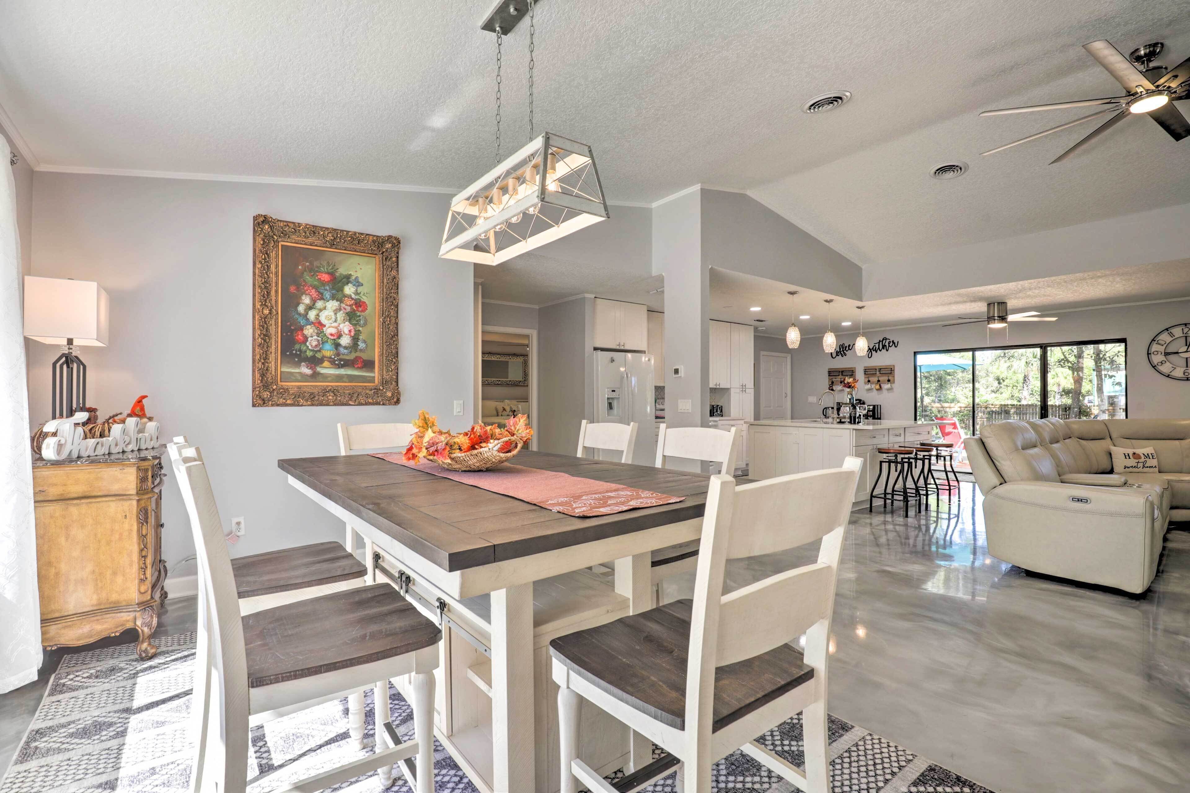 A spacious living area with a dining table, white chairs, a sofa, and a kitchen in the background.