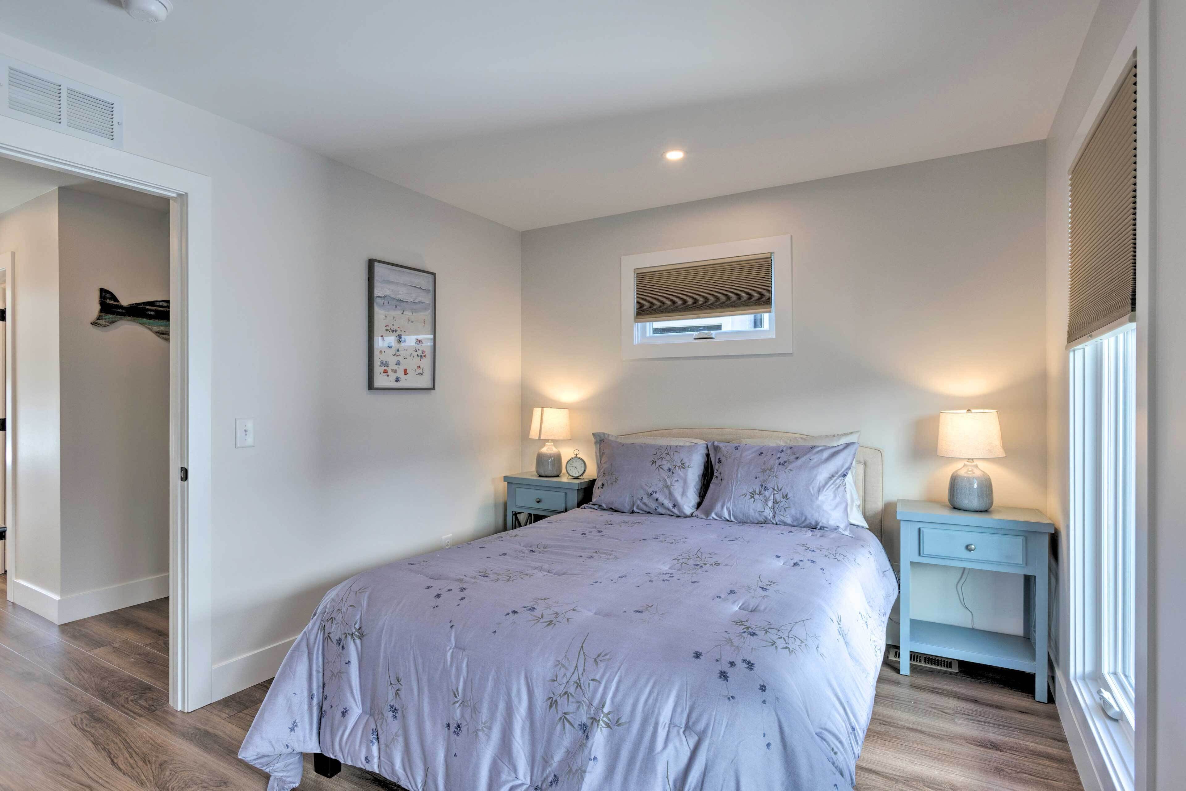 A bedroom with a bed, two bedside tables with lamps, a window with blinds, a door, and a wall-mounted picture.