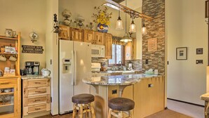 A kitchen with wooden cabinets, a refrigerator, a microwave, and a coffee maker. There are two stools and a hanging light fixture.