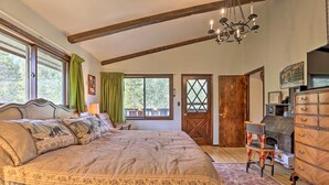 A bedroom with a large bed, wooden beams, a chandelier, a fireplace, and a view of trees through the windows.
