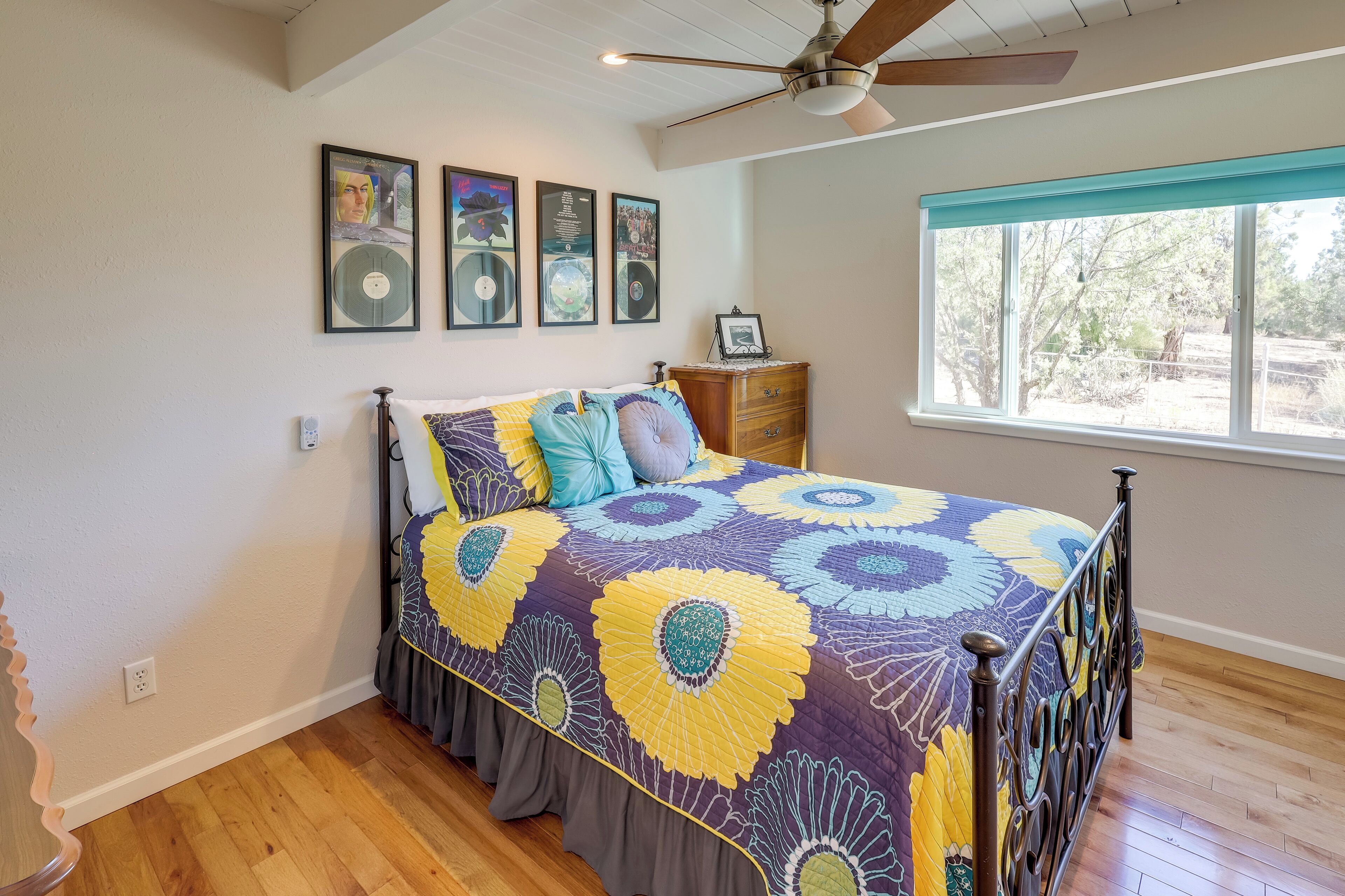 A bedroom with a bed, wooden floor, ceiling fan, window with view, and wall-mounted artwork.