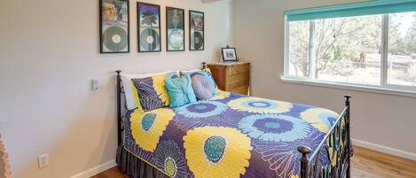 A bedroom with a bed, wooden floor, ceiling fan, window with view, and wall-mounted artwork.