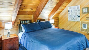 Um quarto com teto de madeira, uma cama com roupa de cama azul, uma mesa de cabeceira com abajur e uma parede com quadros.
