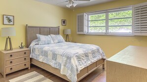 A bedroom with a bed, bedside tables, a ceiling fan, a window with shutters, and a view of greenery outside. - Riviera Beach Home w/ Pool - Walk to Beaches! (Singer Island)
