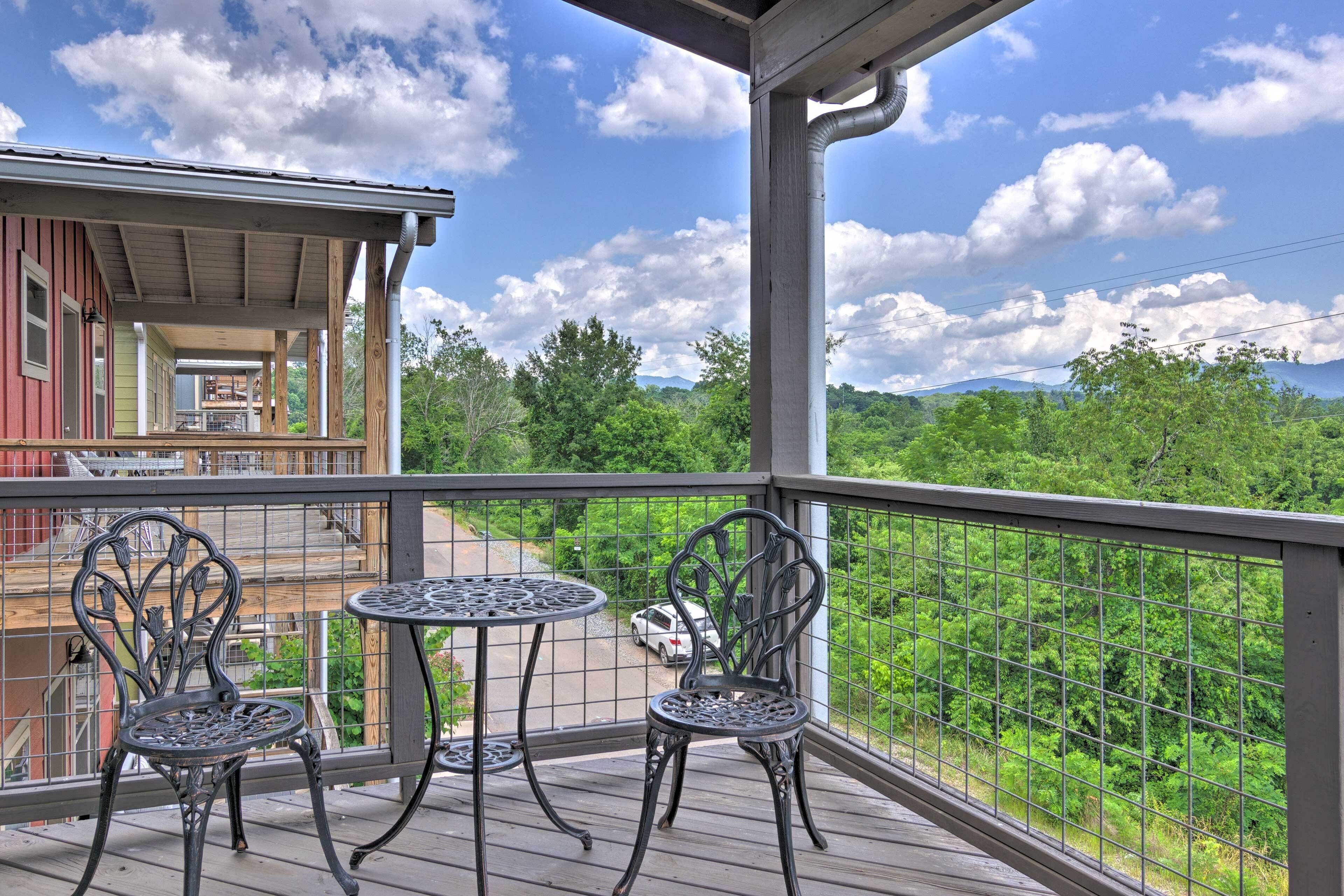 A wooden deck with two metal chairs and a small round table, overlooking a lush green landscape and a distant mountain range.