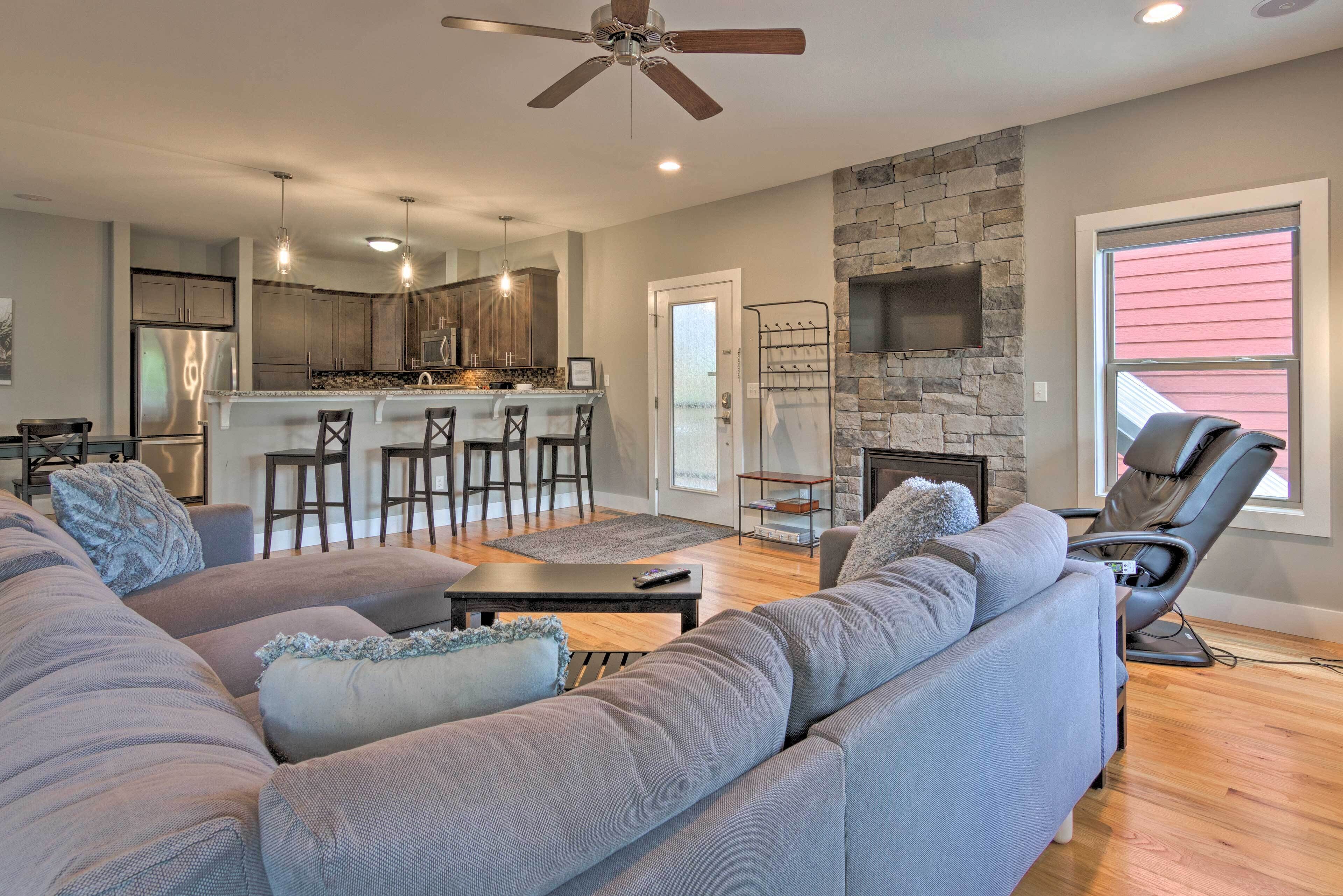 A modern living room with a gray sectional sofa, a stone fireplace, and a bar area in the background.