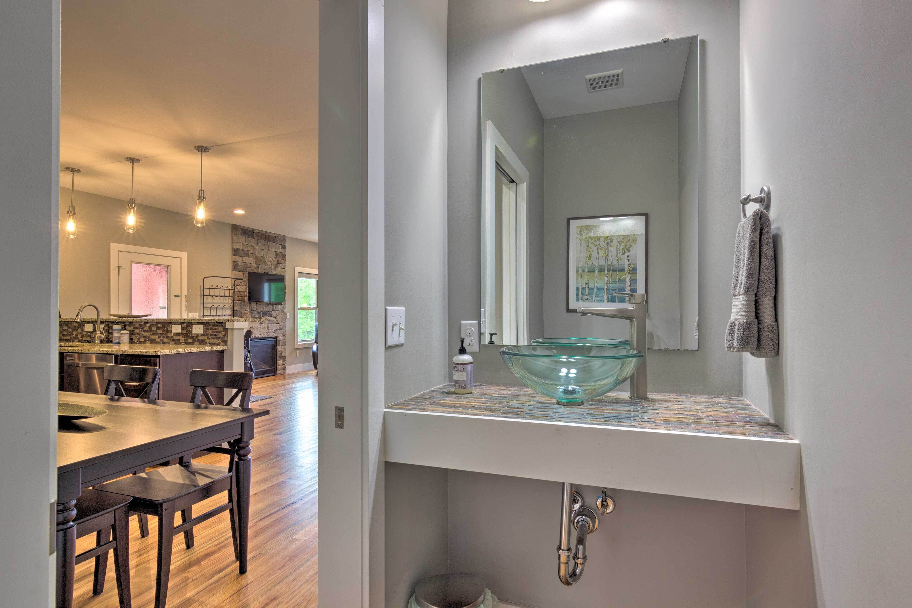 A modern kitchen with a stone backsplash, a dining area with a wooden table and chairs, and a bathroom with a glass bowl sink and a large mirror.