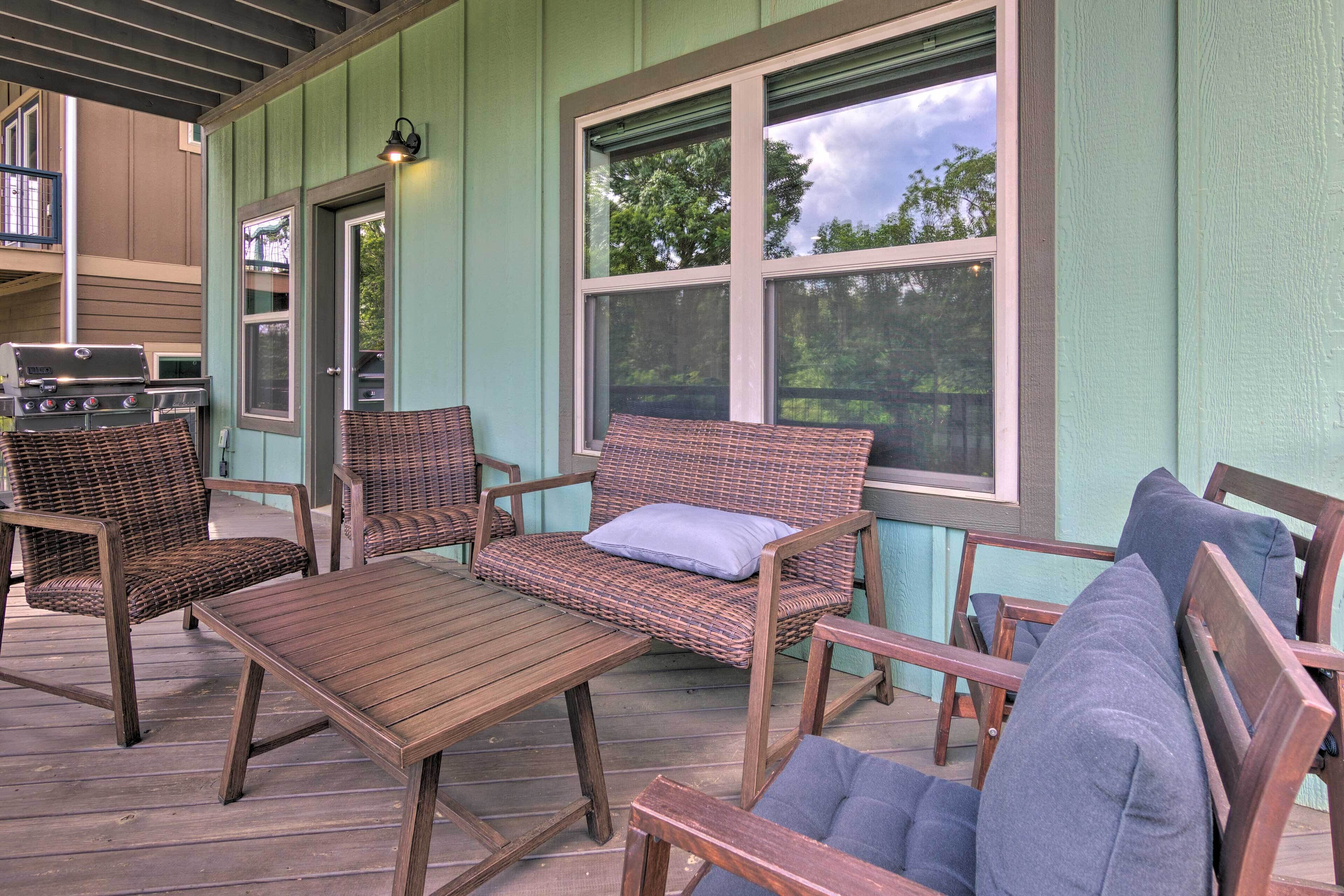A covered outdoor seating area with wicker furniture and a wooden table, a grill, and a view of trees through the windows.