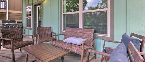 A covered outdoor seating area with wicker furniture and a wooden table, a grill, and a view of trees through the windows.