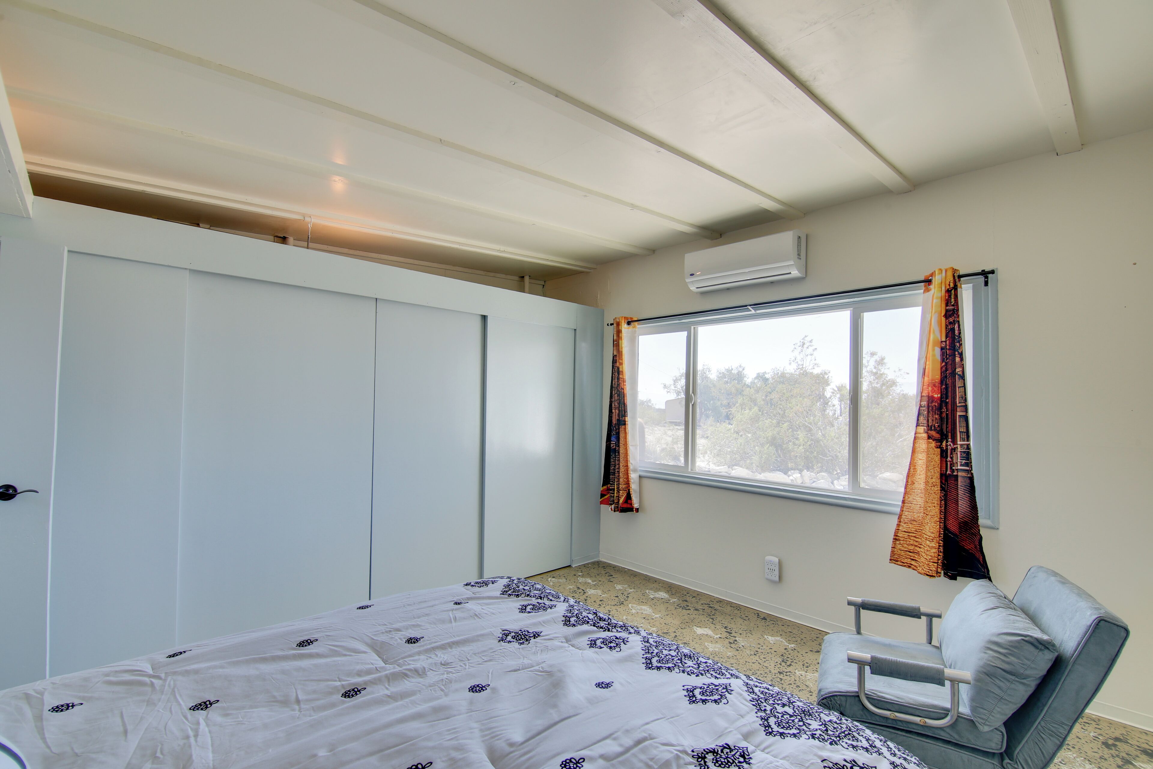 A bedroom with a bed, a chair, a window with curtains, and an air conditioning unit.