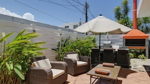 Un patio al aire libre con mobiliario de mimbre, una parrilla y una sombrilla blanca.