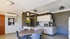 Una cocina compacta con una isla central, armarios blancos y un microondas. Hay una mesa de comedor con sillas y un cartel en la pared.