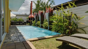 Una piscina con un deck de baldosas de piedra, rodeada de exuberante vegetación y una valla blanca.