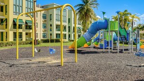 A playground with a slide and swings, surrounded by a building with large windows and palm trees. - Aquatika Resort Condo w/ Private Beach Access! (Loiza)