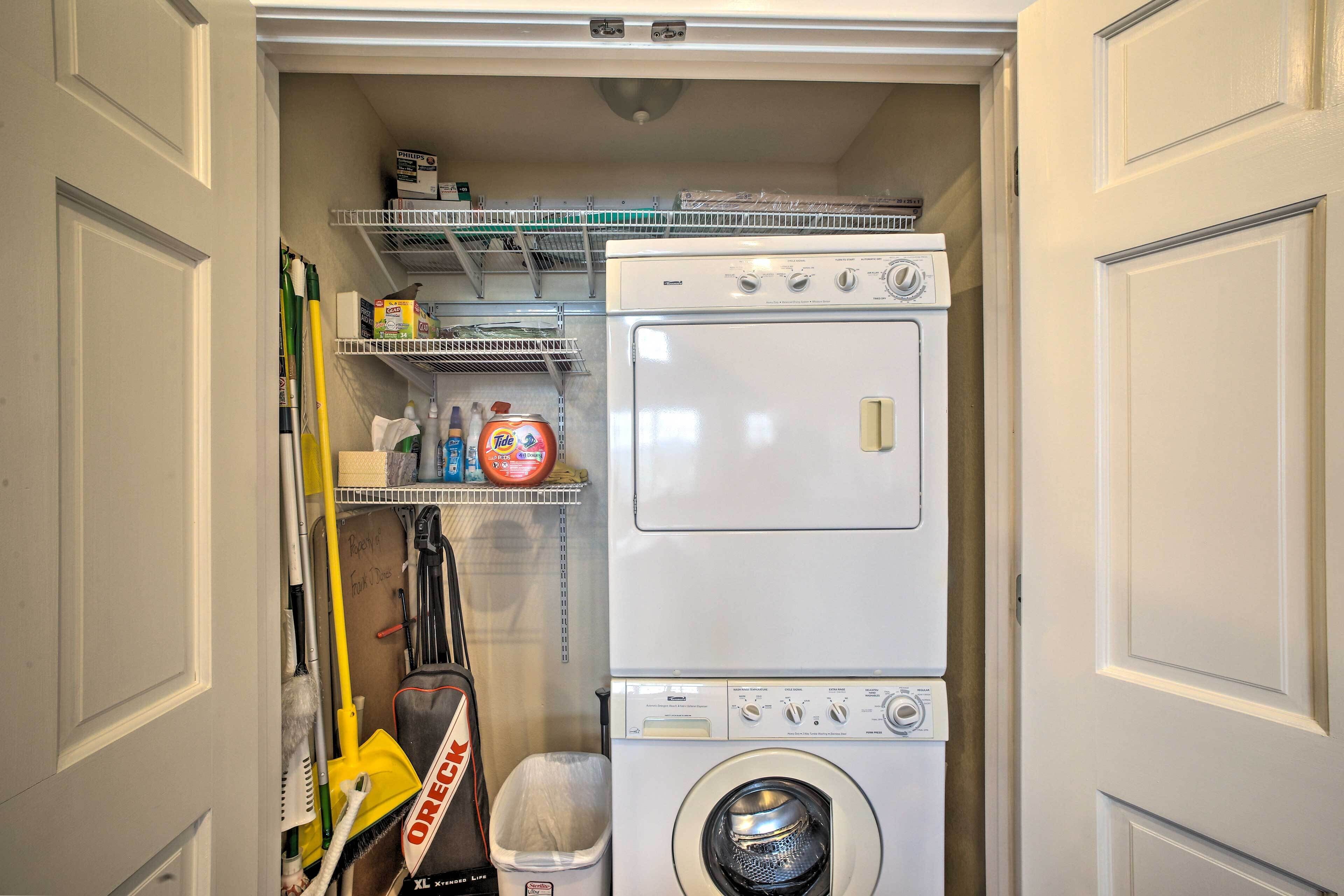A laundry room with a stacked washer and dryer, shelves with cleaning supplies, and a trash bin.