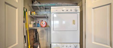 A laundry room with a stacked washer and dryer, shelves with cleaning supplies, and a trash bin.