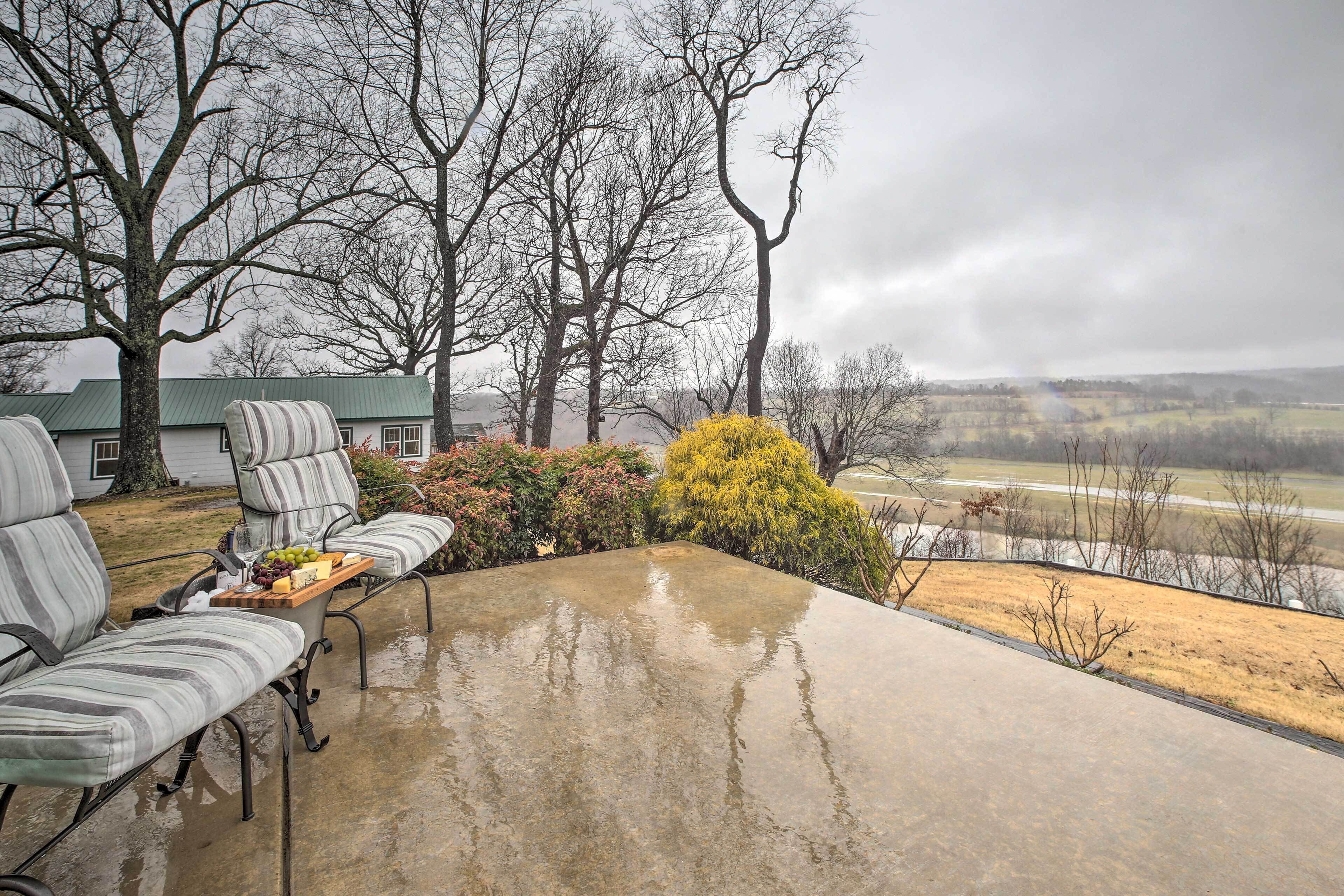 Eine überdachte Terrasse mit Betonboden, zwei gestreifte Stühle, ein Holztisch mit Obstschale und im Hintergrund ein Haus und Bäume.