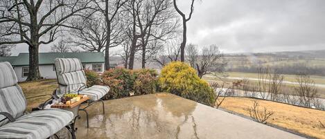 Eine überdachte Terrasse mit Betonboden, zwei gestreifte Stühle, ein Holztisch mit Obstschale und im Hintergrund ein Haus und Bäume.