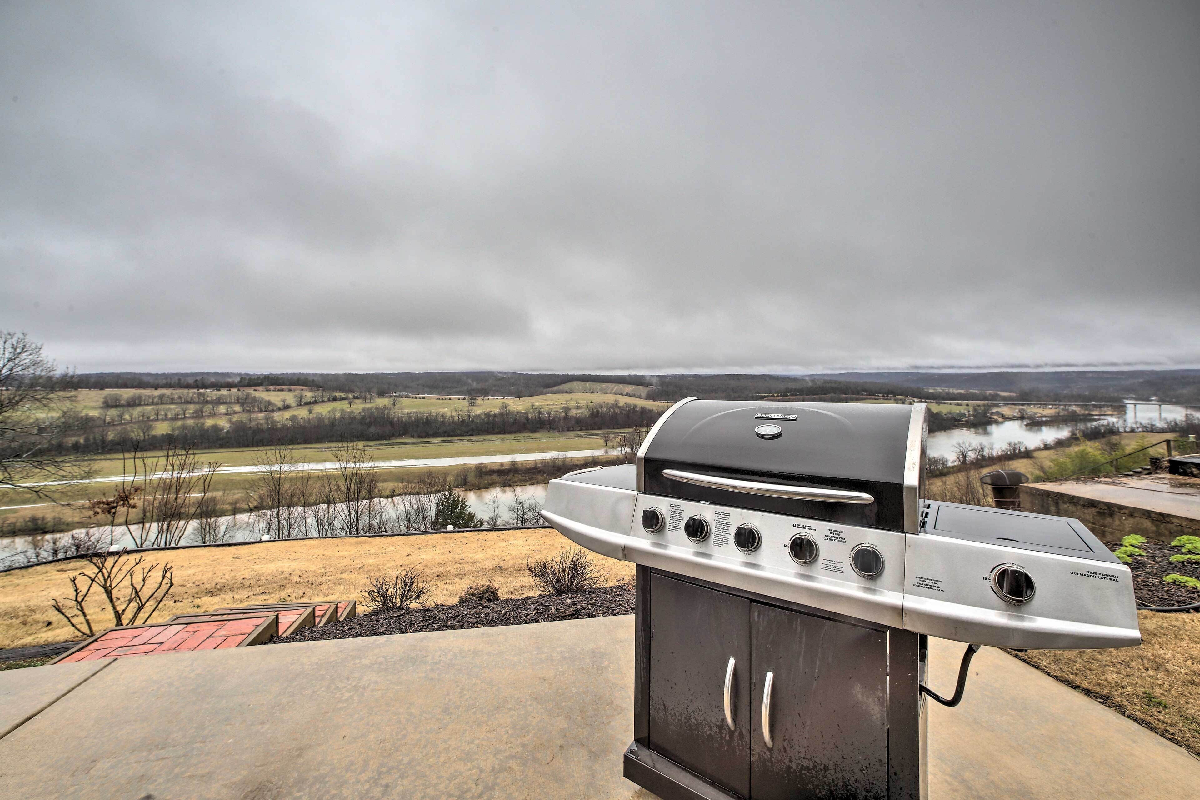 A gas grill with multiple burners and a digital display, set against a backdrop of a river and hilly landscape.