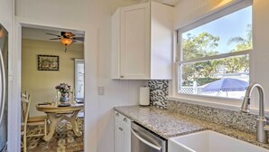 A kitchen with white cabinets, a granite countertop, a stainless steel dishwasher, and a window with a view of trees and a blue umbrella. - Carpinteria Apt w/ Hot Tub - 10 Min Walk to Beach! (Carpinteria)