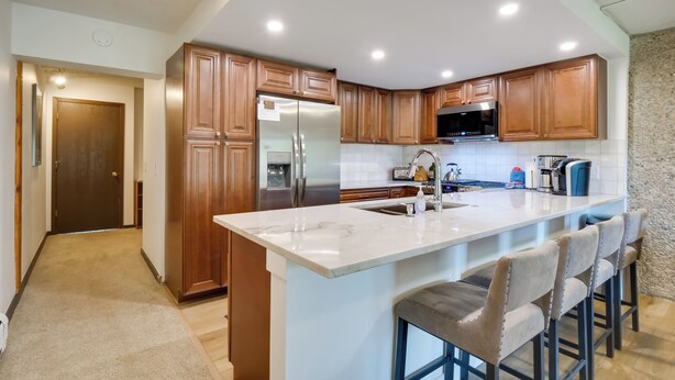 A modern kitchen with wooden cabinets, a stainless steel refrigerator, a microwave, and a marble island with bar stools. - Beautiful Breck Townhome w/ Pool: 1 Mi to Main St! (Breckenridge)