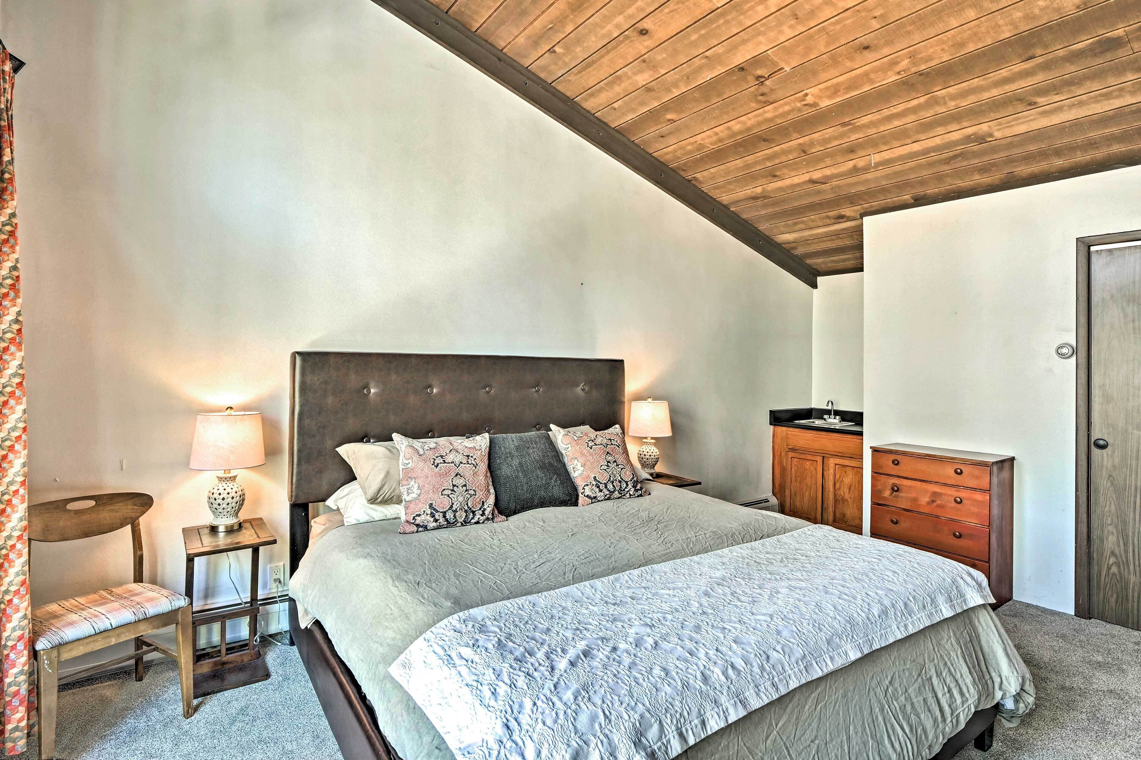 A bedroom with a large bed, two bedside tables with lamps, a wooden headboard, a dresser, and a door.