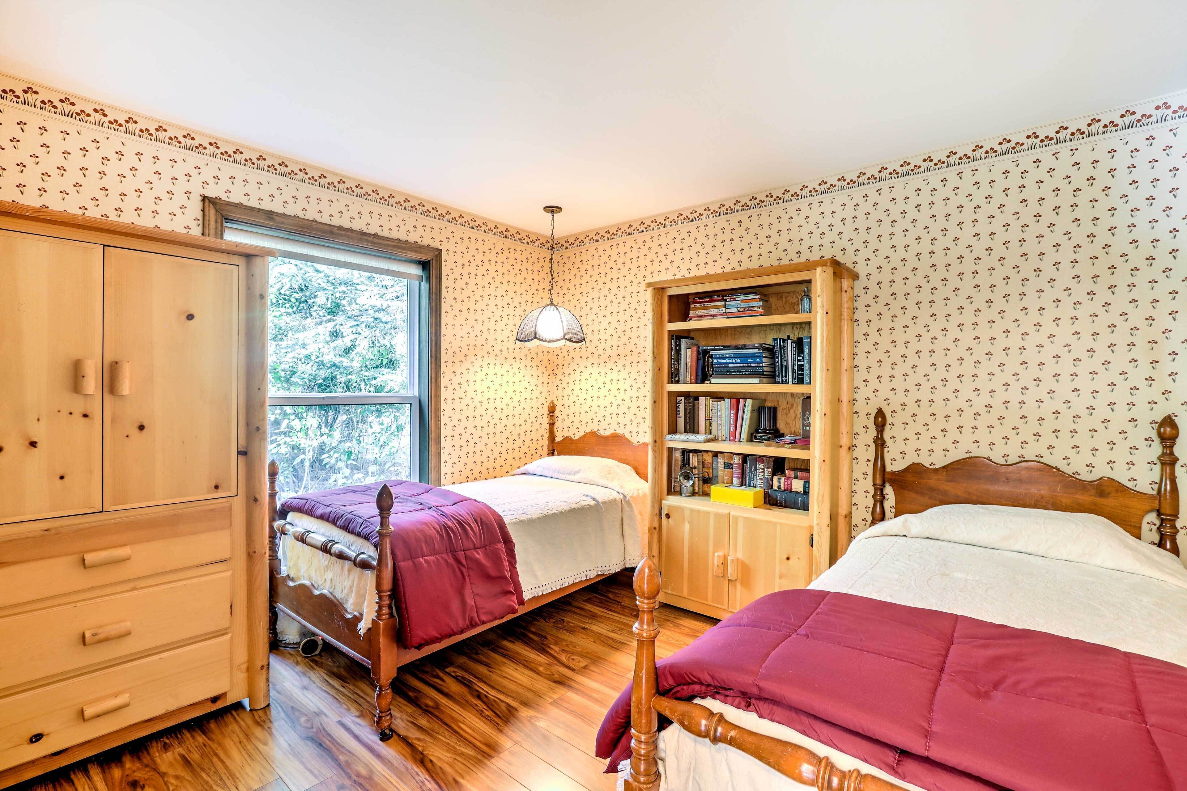 A bedroom with two beds, a wooden wardrobe, a bookshelf, and a window with a view of greenery.
