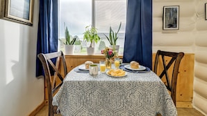 Una mesa de comedor con mantel estampado, rodeada de sillas de madera, con vistas al exterior, plantas en macetas y alimentos para el desayuno.