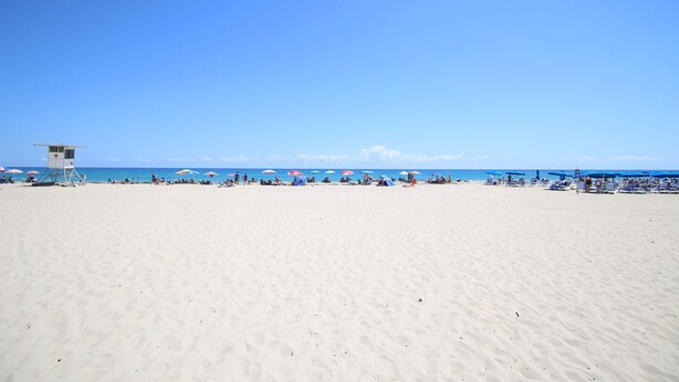 Ein weiter Sandstrand mit einem Rettungsturm und Sonnenschirmen.